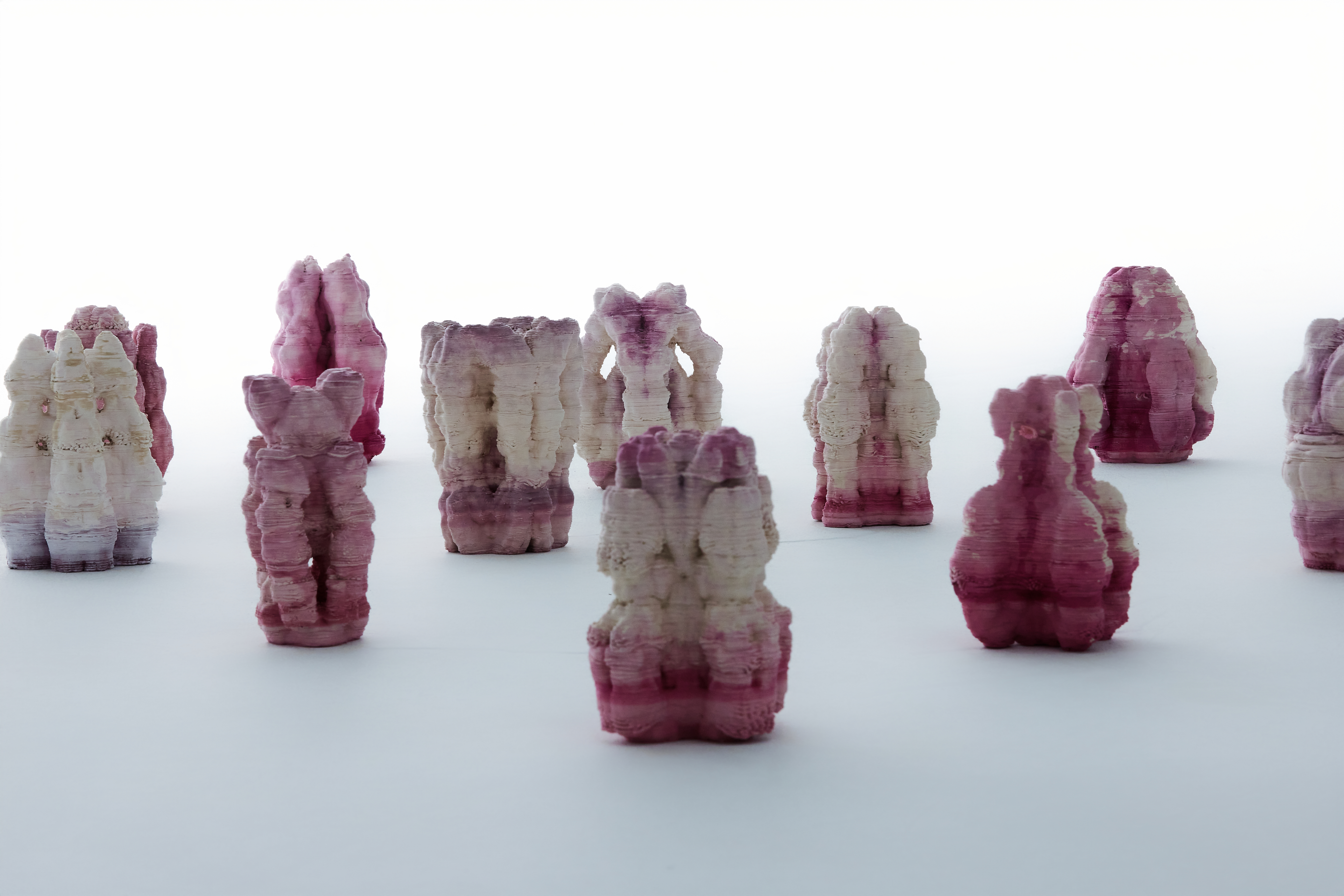 The fourteen ceramic sculptures of Bactereature arranged on a white ground, each carrying an uneven pink-to-magenta gradient left by bacterial growth.