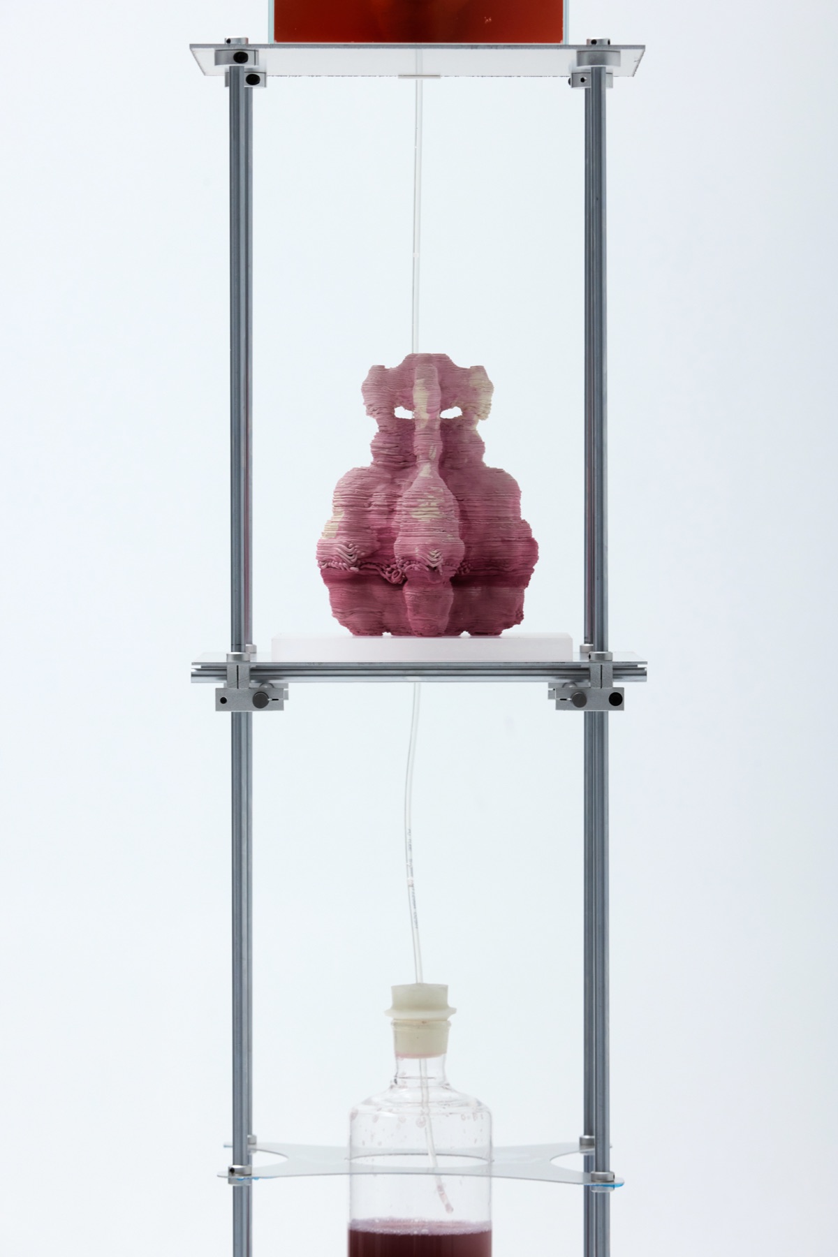 A stained ceramic piece on a white block on an aluminium-frame plinth, a glass reservoir of pigment medium suspended on the shelf below, connected by tubing.