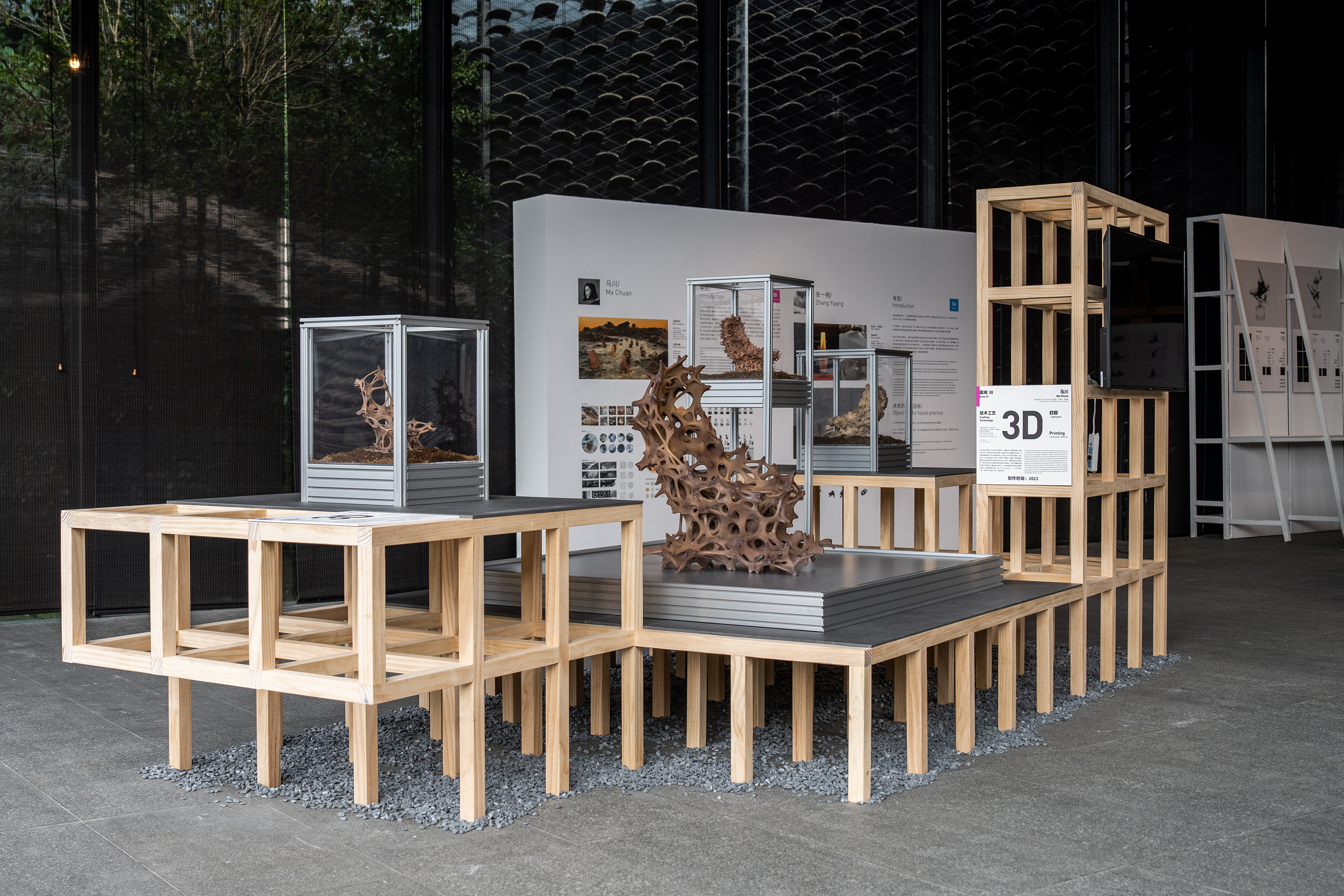 Installation view of Enso at the China Folk Art Museum: wood-frame plinths on grey gravel carrying glass vitrines, each holding a brown lattice sculpture, with a printed wall panel behind.